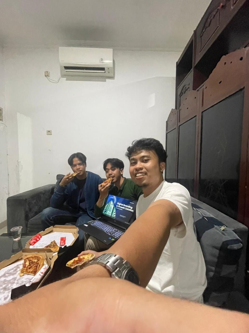 Swafoto tiga anggota tim makan pizza di sofa, dengan laptop terbuka di depan mereka.